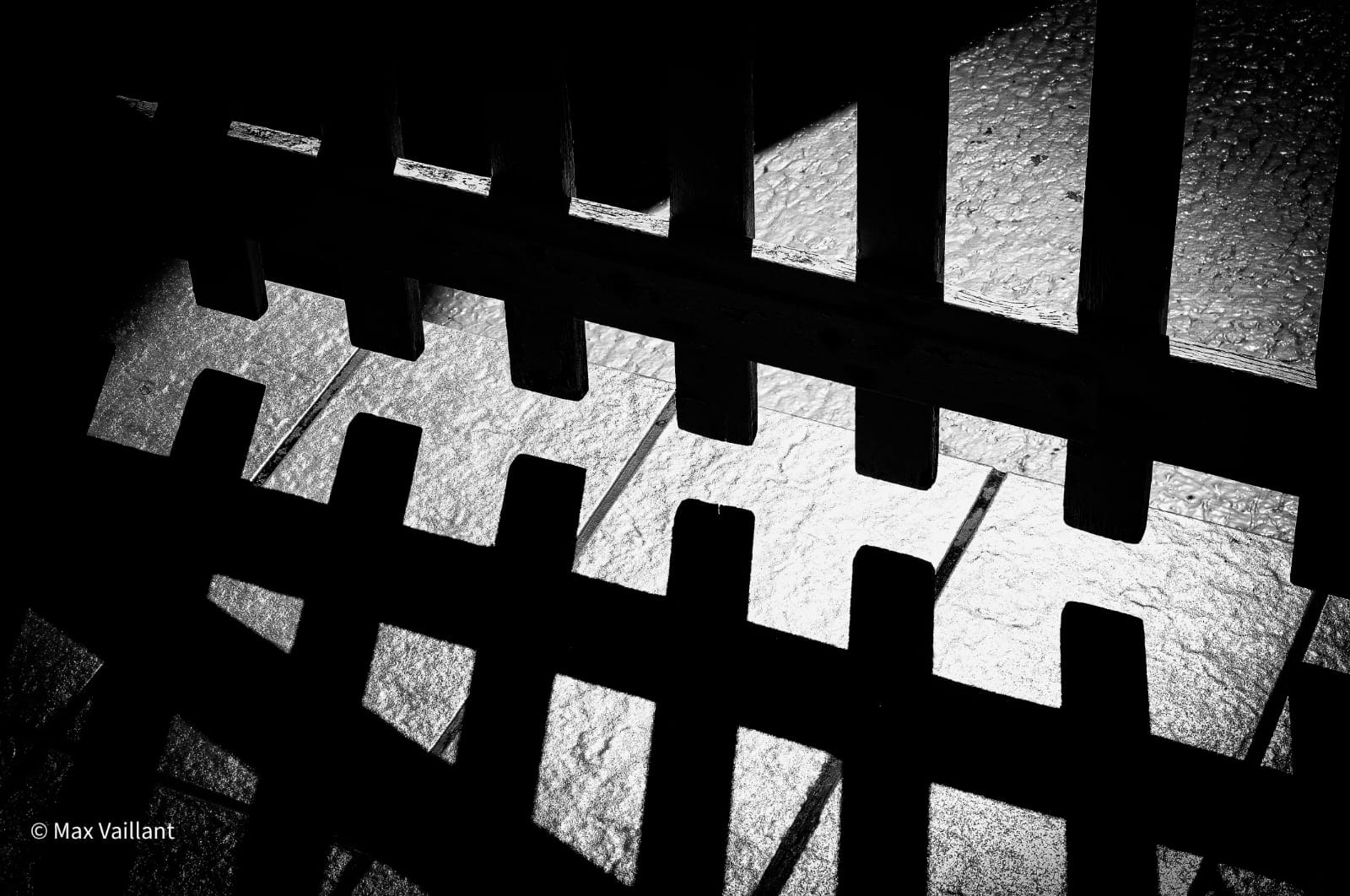 Ombre portée d'une grille de bois, en diagonale sur un sol de pierre clair et grenu.
